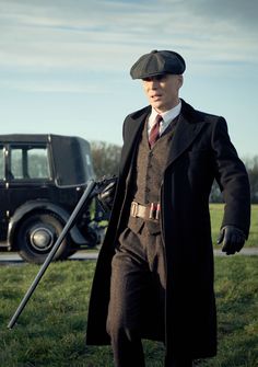 a man in a suit and hat holding a cane while standing next to an old car