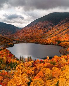 a lake surrounded by trees in the middle of mountains with fall foliage around it and cloudy skies above