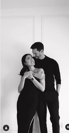 a man and woman standing next to each other with their arms around each other in front of a white wall