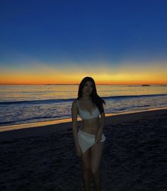 a woman standing on top of a beach next to the ocean at sunset with her hands in her pockets