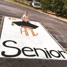 a man sitting on the ground in front of a sign that says senior with an orange and black graduation cap