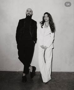 a man and woman standing next to each other in front of a white wall wearing black clothes