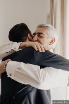 two men are hugging each other in front of a window and one man is wearing a suit