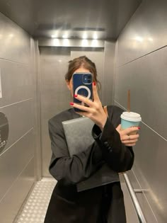 a woman taking a selfie with her cell phone in an elevator holding a cup of coffee