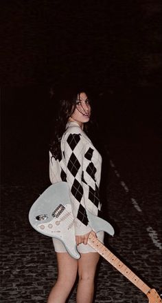 a woman holding an electric guitar in her right hand and posing on the street at night