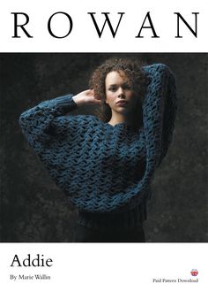 a woman with curly hair wearing a blue knitted sweater and black pants, standing in front of a dark background