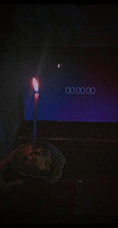 a person holding a cupcake with a lit candle on it in front of a laptop