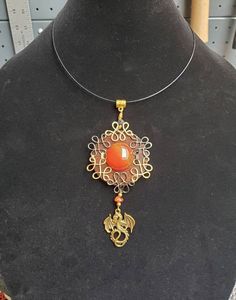 This mighty fantasy necklace embraces the majesty, beauty, and power of dragons. Wear anyplace that you would like to portrait beauty and power.   A fiery red carnelian acts as the centerpiece to the overall fantasy design. It is surrounded by a dragon hoard of golden knotwork that alternates with some fire singed looking hematite toned knotwork (both made utilising tarnish resistant Parawire). A Small lava stone bead sits above while a gold toned dragon charm and orange agate piece hang below. Before the wire wrapping and accents were added, the stained wood underwent a protective treatment process. First, layers of a hypoallergenic shellac were added. It was then further treated with layers of spar urethane to protect the wood from sunlight and moisture. (This does not mean that I ever r Red Carnelian Spiritual Jewelry, Red Hand Wrapped Necklace As Gift, Hand Wrapped Red Necklace As Gift, Red Cabochon Spiritual Jewelry, Spiritual Red Cabochon Jewelry, Fantasy Wire Wrapped Jewelry Gift, Fantasy Wire Wrapped Jewelry As Gift, Fantasy Style Wire Wrapped Jewelry Gift, Adjustable Red Fantasy Jewelry