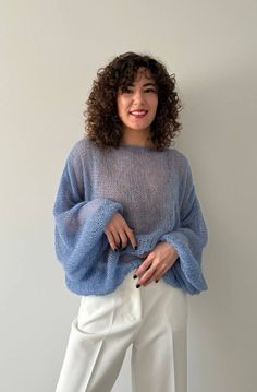 a woman standing in front of a white wall wearing a blue sweater and white pants