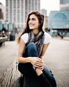 a woman sitting on top of a wooden bench in front of tall buildings with her arms crossed