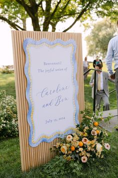 a sign that is sitting in the grass with flowers on it and people standing around