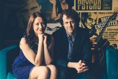 a man and woman sitting on a blue couch in front of a wall with posters