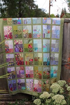 a quilt is hanging on the side of a fence next to some flowers and plants