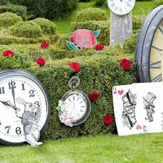 several clocks sitting on top of a lush green field next to bushes and red roses