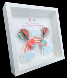 a white shadow box with some paper cut outs in the shape of a rainbow and clouds