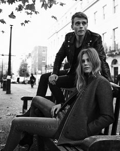 two people sitting on a bench in the middle of a city street, one man is leaning against the back of another woman