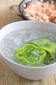 a white bowl filled with ice and green peppers on top of a wooden table next to a frying pan