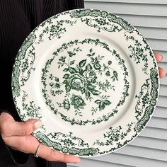 a person holding a green and white plate with flowers on the rim, in front of a window