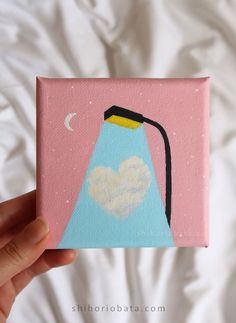 a hand holding up a small painting with a light on it's side and a moon in the sky