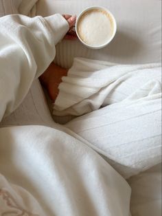 someone is holding a cup of coffee in their hand while laying in bed with white sheets