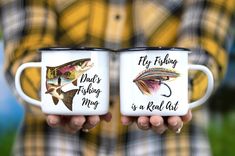 two coffee mugs with fishing designs on them are held in front of each other