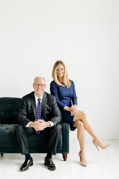 a man and woman sitting on a couch in front of a white wall, posing for the camera