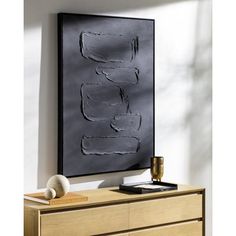 a black piece of art hangs on the wall above a dresser with a vase and other items