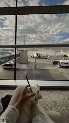 a person sitting in front of an airport window holding a cup with a straw and looking out at planes on the tarmac