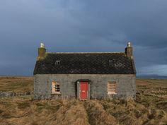 an old stone house with a red door in the middle of a grassy field under a cloudy sky