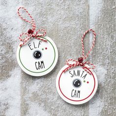 two ceramic ornaments with the words elf and santa's cam hanging from twine