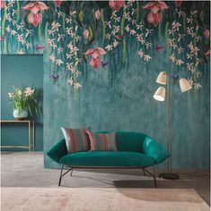 a green couch sitting in front of a wall with pink flowers on it and a lamp next to it