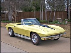 a yellow corvette sits parked in front of a fenced area with trees and bushes