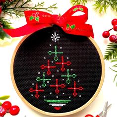 a cross stitch christmas tree ornament with red ribbon and holly wreath around it