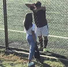 two people standing on the side of a tennis court near a fence and grass area