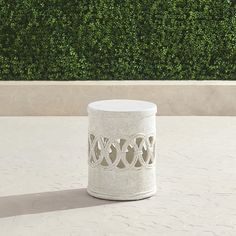 a white vase sitting on top of a table next to a green hedge covered wall