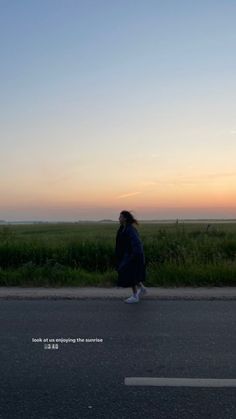 a woman walking down the street in front of a field at sunset or dawn with her back turned to the camera