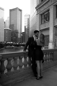 a man and woman leaning against a railing in front of a body of water with skyscrapers behind them