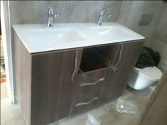 a bathroom vanity with two sinks and a toilet in the back ground, next to a wall mounted trash can