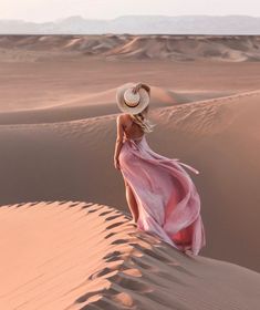 a woman in a pink dress and hat walking through the sand dunes with her back to the camera