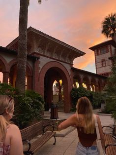 two women walking towards a building with palm trees in the foreground and pink clouds in the background