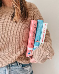 a woman is holding three books in her hands while wearing a sweater and denim shorts