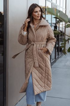 a woman standing on the sidewalk wearing a tan coat and blue jeans with her hands in her pockets