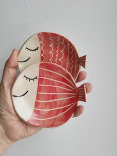 a hand holding a red and white ceramic fish bowl with eyes closed on it's side