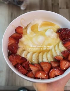 a person holding a bowl of food with strawberries and bananas in it on top of a wooden table