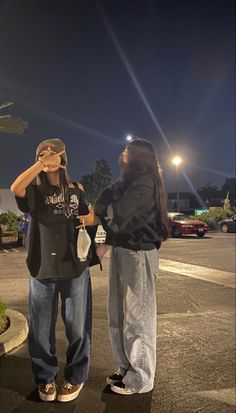 two people standing in the middle of a parking lot at night with their hands to their eyes