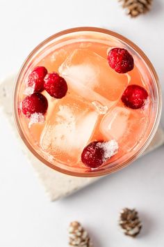 a drink with ice, raspberries and pine cones on the table next to it