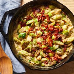 a skillet filled with vegetables and bacon on top of a wooden table next to a spoon