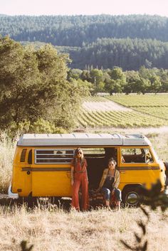 two women sitting in the back of a yellow van on top of a grass covered field