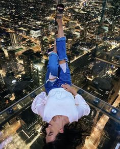 a woman hanging upside down on the glass floor of a high rise building at night