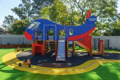 An outdoor childcare playground with a large blue and red airplane with slides in the middle of the playground with a yellow cartoon looking road around it. Around the road there is grass. Montessori Education, Childcare, Montessori, Preschool